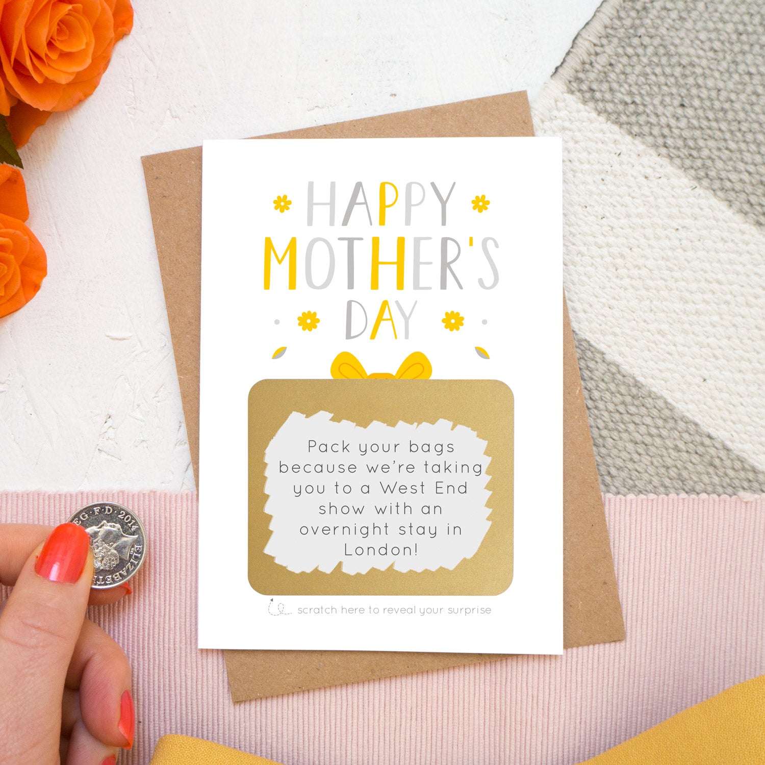 A mother’s day present scratch card lying on a kraft brown envelope on top of a white, grey and pink background with orange roses poking in the top left. The card reads ‘Happy Mother’s day’ and the gold present has been scratched off  with a coin to reveal a custom message to a mum. This card is in the yellow and grey colour palette.