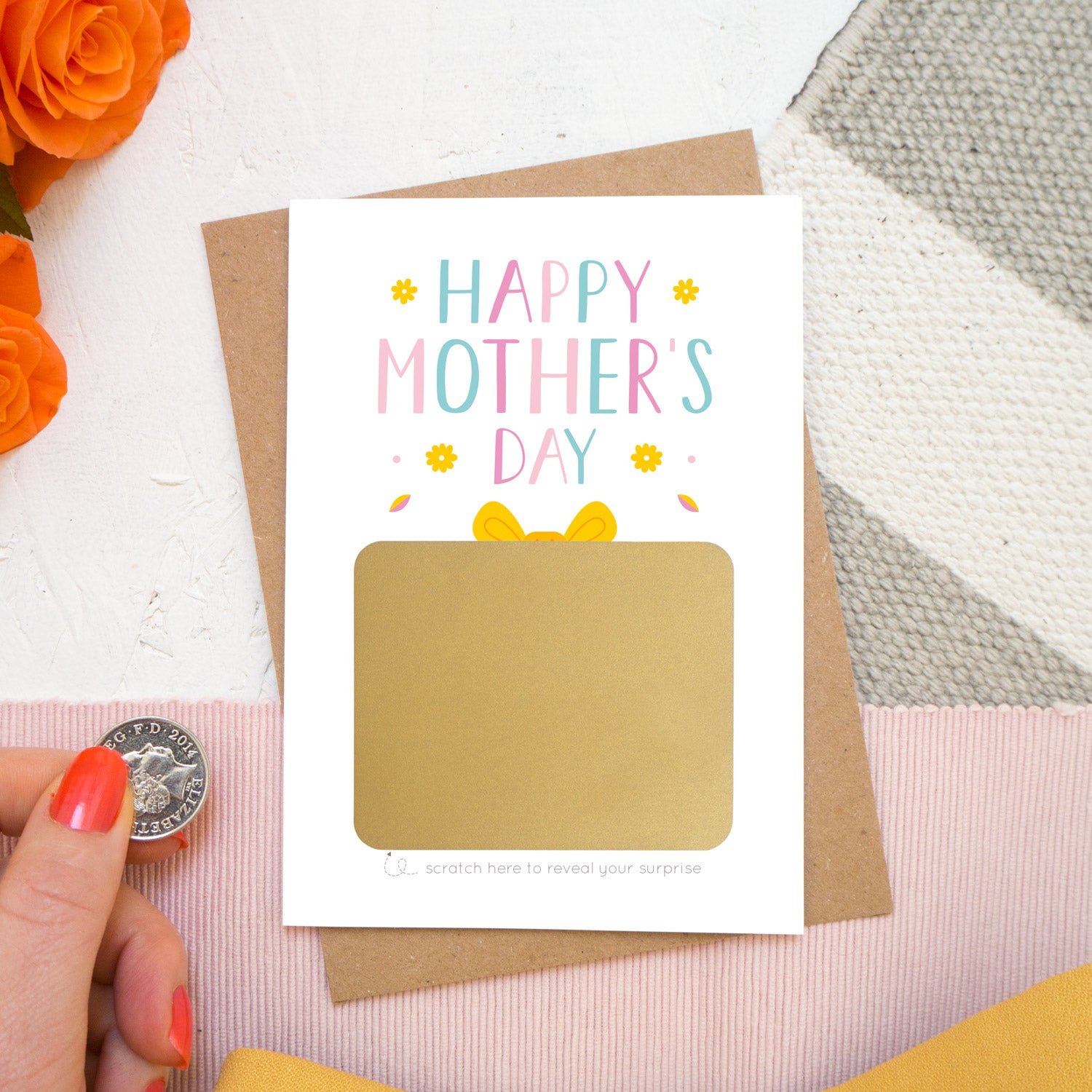 A mother’s day present scratch card lying on a kraft brown envelope on top of a white, grey and pink background with orange roses poking in the top left. The card reads ‘Happy Mother’s day’ and the gold present is yet to be scratched off. This card is in the pink and blue colour palette.