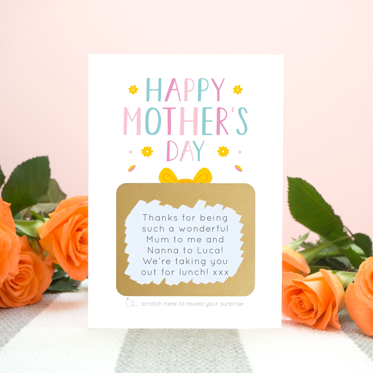 A mother’s day present scratch card standing in front of orange roses on a grey and white striped rug against a pink background. The card reads ‘Happy Mother’s day’ and the gold present has been scratched off to reveal a custom message to a mum. This card is in the pink and blue colour palette.