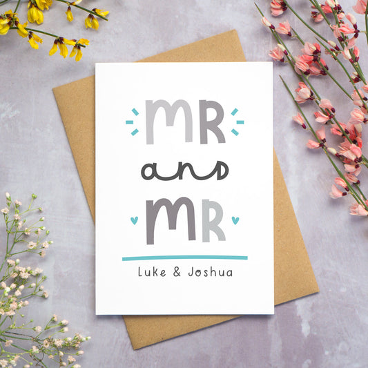 A Mr and Mr personalised wedding congratulations card lying flat on top of a kraft brown envelope on a grey background with yellow, pink and white foliage coming in from the sides. This is the grey and blue version of the card.