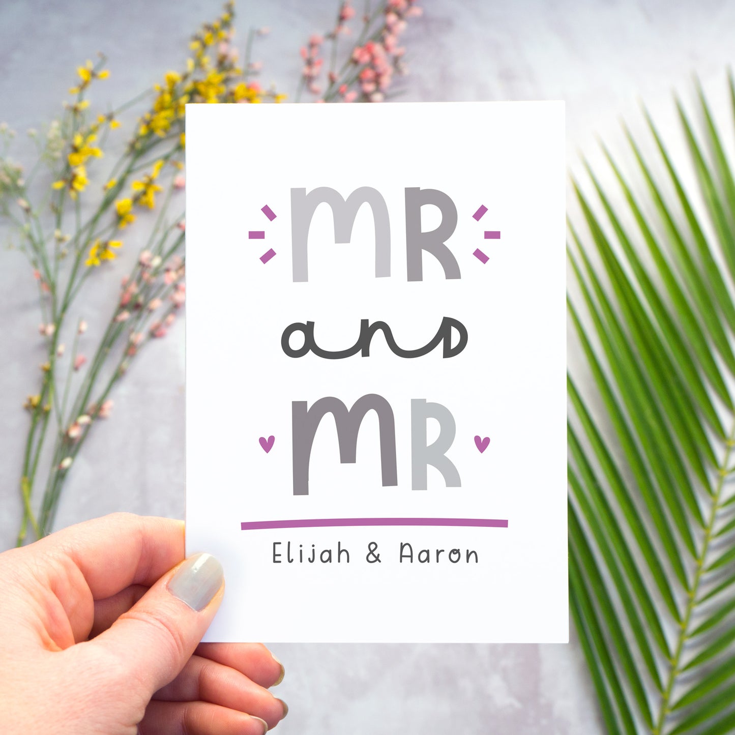 A Mr and Mr personalised wedding congratulations card being held over a grey back drop with larger pieces of green, pink and yellow foliage blurred out in the background. This is the grey and purple version of the card.