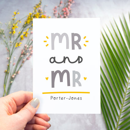 A Mr and Mr personalised wedding congratulations card being held over a grey back drop with larger pieces of green, pink and yellow foliage blurred out in the background. This is the grey and yellow version of the card.