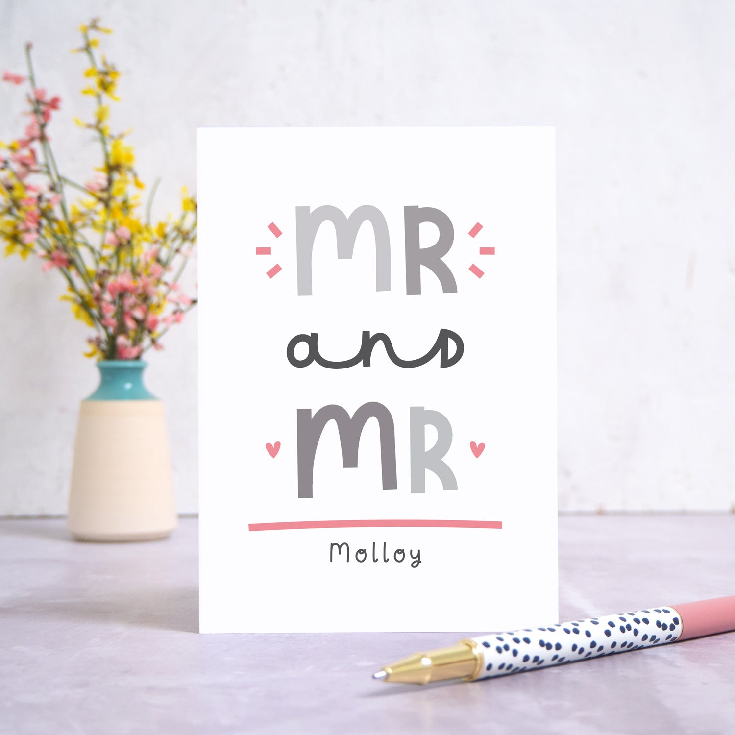 A Mr and Mr personalised wedding congratulations card stood in front of a white background and a small vase of yellow and pink flowers. There is a white and black spotty pen in the foreground. This is the grey and pink version of the card.