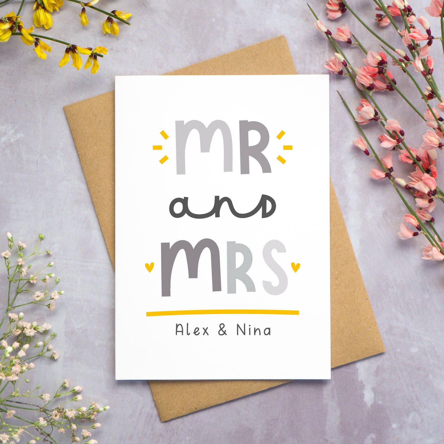 A Mr and Mrs personalised wedding congratulations card lying flat on top of a kraft brown envelope on a grey background with yellow, pink and white foliage coming in from the sides. This is the grey and yellow version of the card.