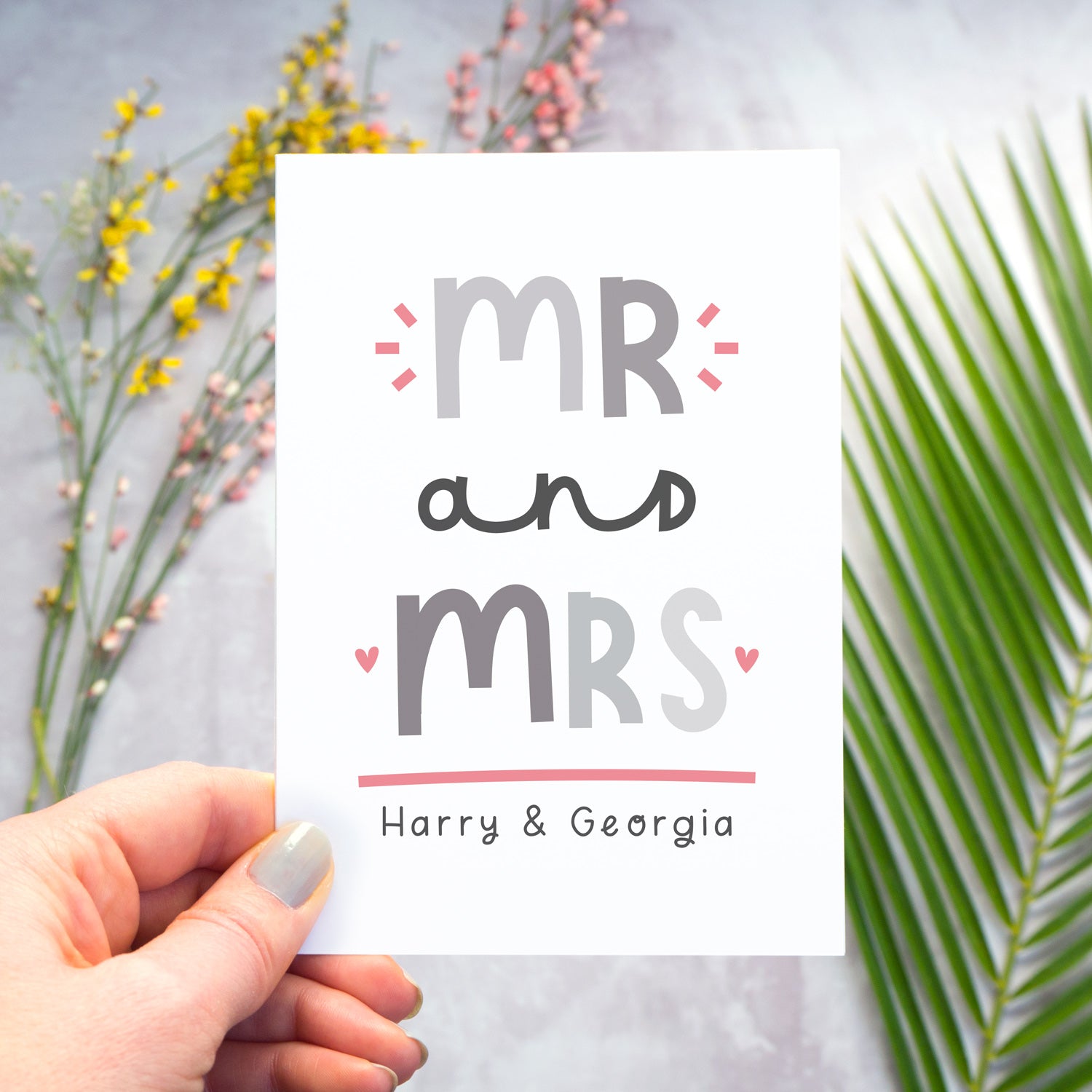 A Mr and Mrs personalised wedding congratulations card being held over a grey back drop with larger pieces of green, pink and yellow foliage blurred out in the background. This is the grey and pink version of the card.