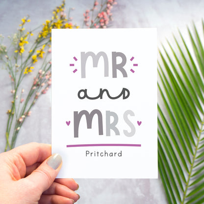 A Mr and Mrs personalised wedding congratulations card being held over a grey back drop with larger pieces of green, pink and yellow foliage blurred out in the background. This is the grey and purple version of the card.