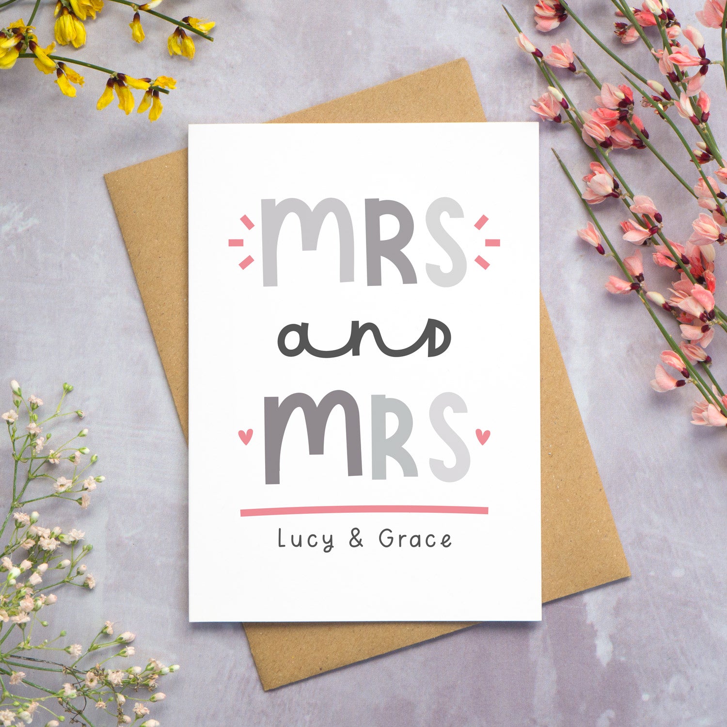 A Mrs and Mrs personalised wedding congratulations card lying flat on top of a kraft brown envelope on a grey background with yellow, pink and white foliage coming in from the sides. This is the grey and pink version of the card.