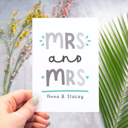 A Mrs and Mrs personalised wedding congratulations card being held over a grey back drop with larger pieces of green, pink and yellow foliage blurred out in the background. This is the grey and blue version of the card.