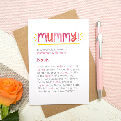 A personalised Mummy dictionary definition card lying flat on top of a kraft brown envelope on top of a pink runner and a grey and white striped rug. There is an orange rose in the bottom left corner and a pen for scale on the right. The card features hand drawn writing in varying tones of pink and a definition of what a mummy is with key words highlighted in pink.