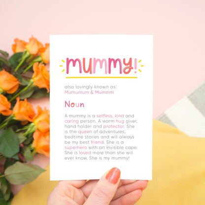 A personalised Mummy dictionary definition card being held over a pink background with a grey and white striped rug, yellow skirt and a bunch of orange roses to the left. The card features hand drawn writing in varying tones of pink and a definition of what a mummy is with key words highlighted in pink.