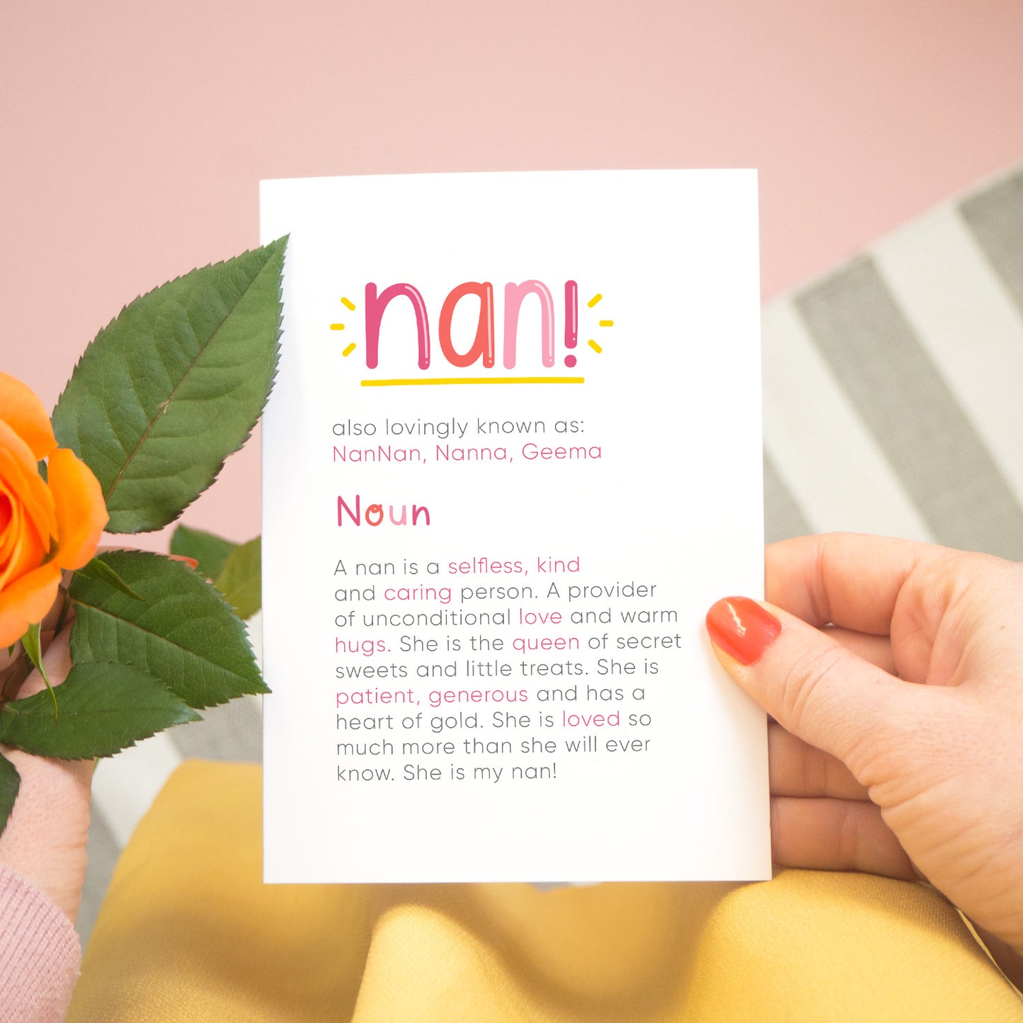 A personalised Nan dictionary definition card being held over a pink background with a grey and white striped rug, yellow skirt and a single orange rose to the left. The card features hand drawn writing in varying tones of pink and a definition of what a nan  is with key words highlighted in pink.