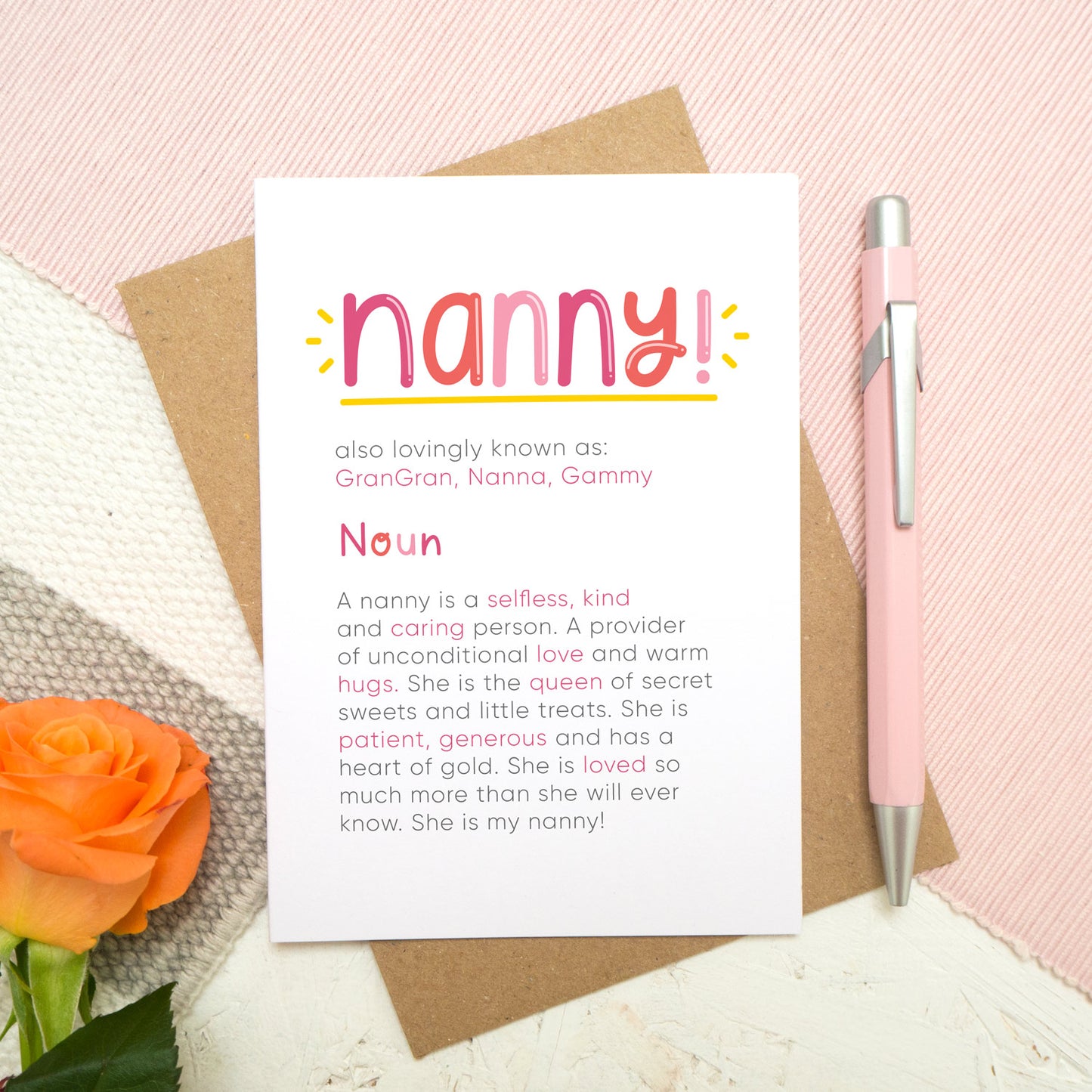 A personalised Nanny dictionary definition card lying flat on top of a kraft brown envelope on top of a pink runner and a grey and white striped rug. There is an orange rose in the bottom left corner and a pen for scale on the right. The card features hand drawn writing in varying tones of pink and a definition of what a nanny is with key words highlighted in pink.