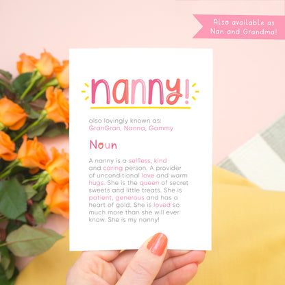 A personalised Nanny dictionary definition card being held over a pink background with a grey and white striped rug, yellow skirt and a bunch of orange roses to the left. The card features hand drawn writing in varying tones of pink and a definition of what a nanny is with key words highlighted in pink.