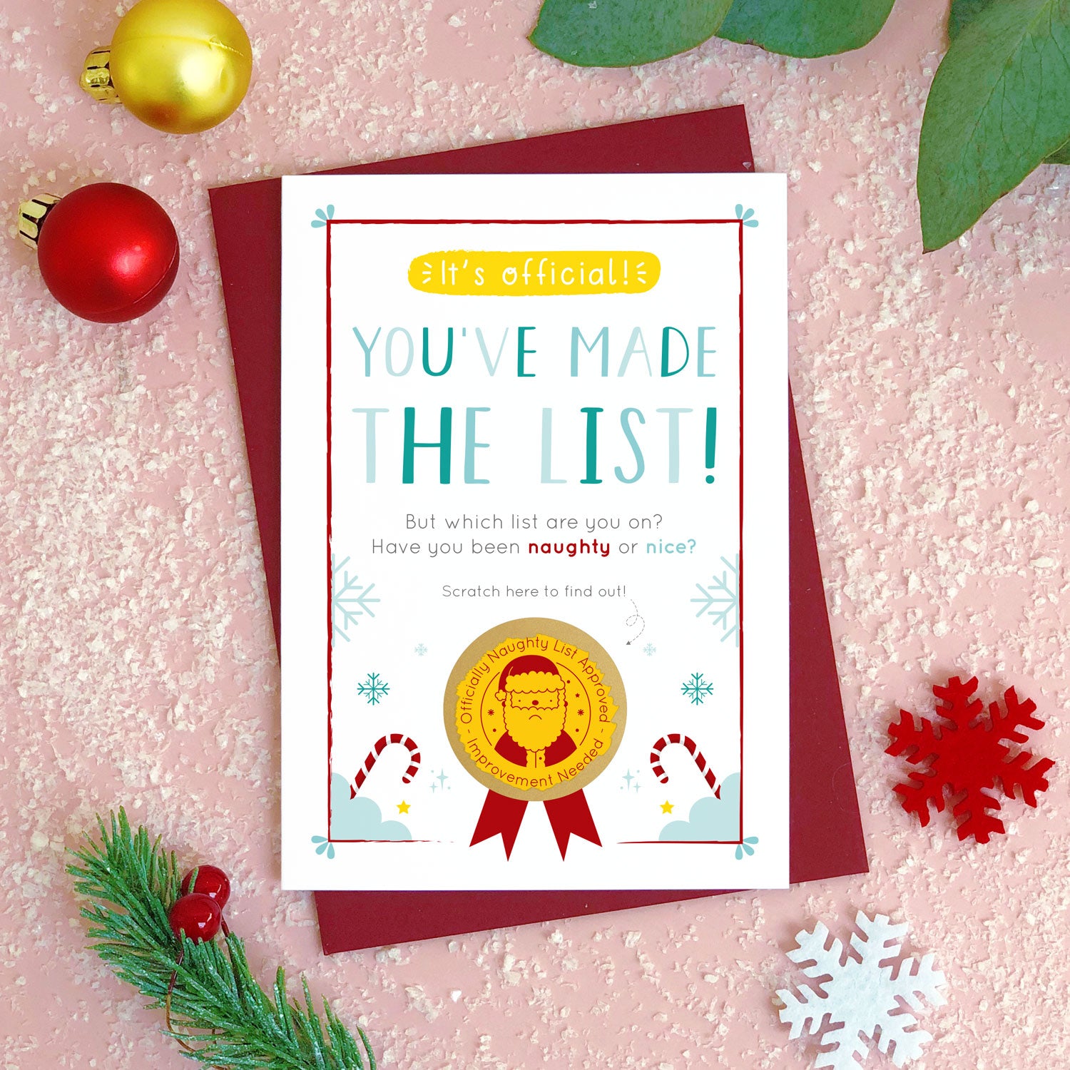 A naughty or nice christmas scratch card photographed flat lying on a red wine coloured envelope on a pink surface surrounded by fake snow, baubles and foliage. The gold panel has been completely scratched away to reveal the sad santa and naughty list approved stamp!