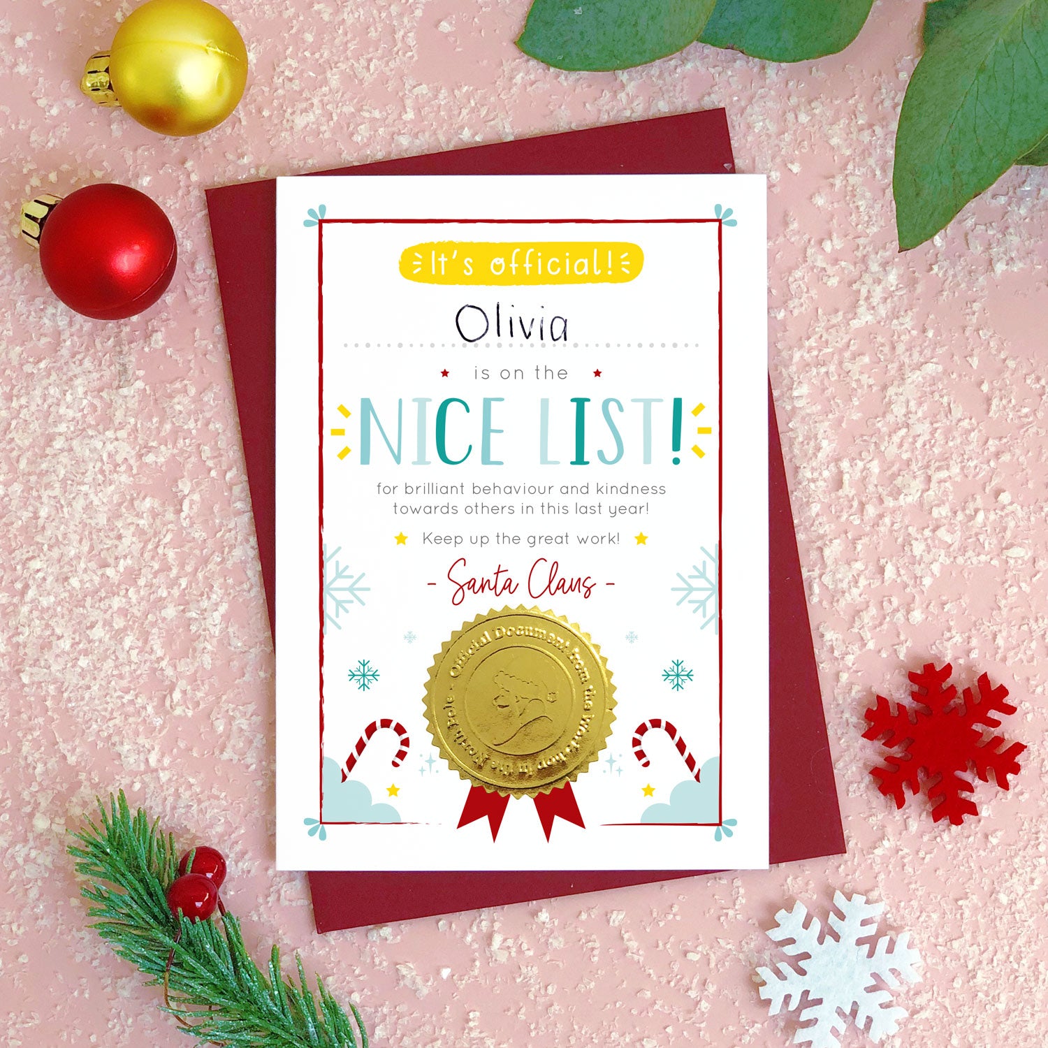 A nice list certificate card photographed flat lay style on top of a pink background covered in ‘snow’, pieces of foliage, snowflakes and a bauble. The card has a blank space for you to hand write a childs name and in this example a biro has been used to show this. It also features a shiny gold seal with the face of a santa character.