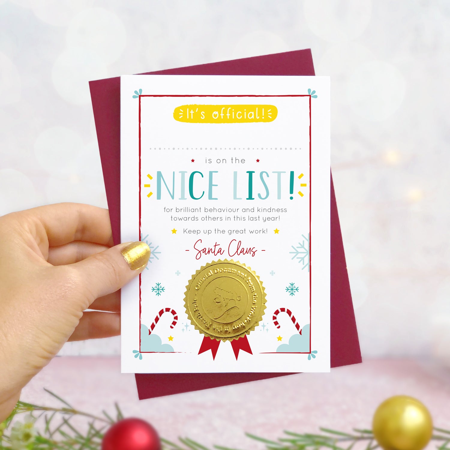 A nice list certificate card photographed in hand in front of a grey and dusty pink background with foliage and baubles in the foreground. The card has a blank space for you to write a childs name. It also features a shiny gold seal with the face of a santa character.