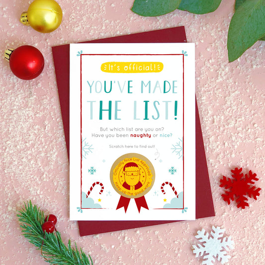 A naughty or nice christmas scratch card photographed flat lying on a red wine coloured envelope on a pink surface surrounded by fake snow, baubles and foliage. The gold panel has been completely scratched away to reveal the happy santa and good list approved stamp!