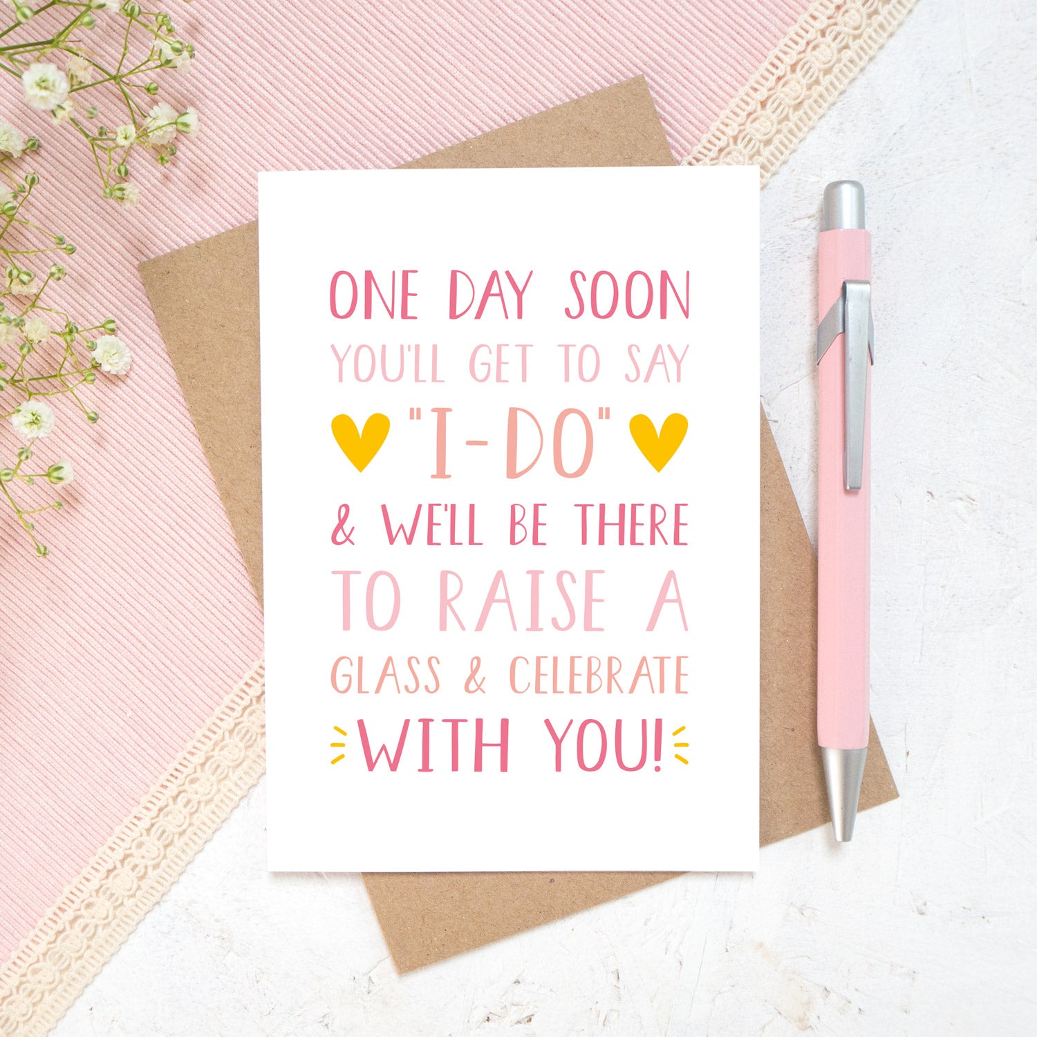 One day soon postponed wedding card in tones of pink. Photographed on a white and pink background with a hint of small flowers and a pen.