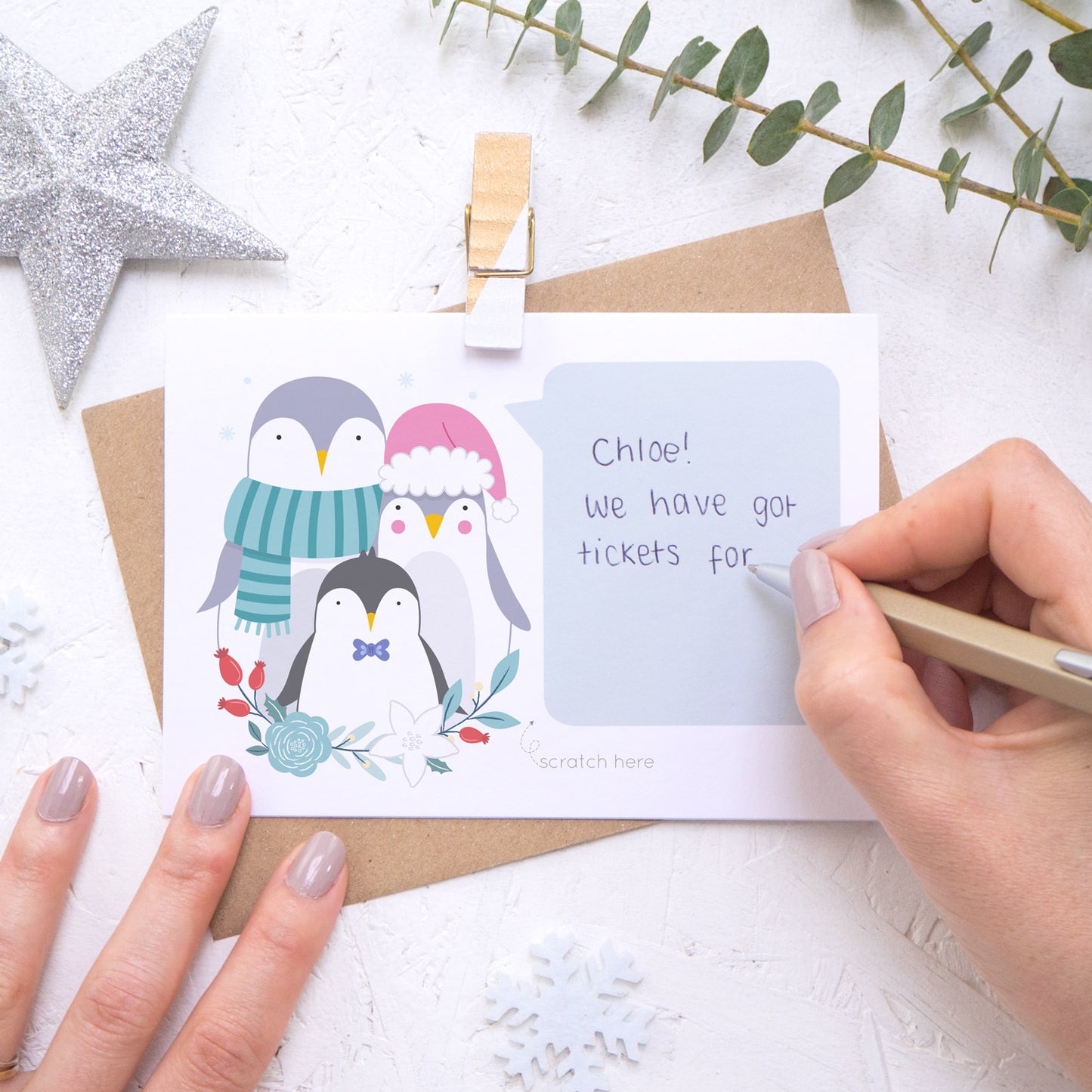 A personalised penguin family scratch card showing where to write the hand written message. Shot on a white background with grey and green festive props and a glittery star.