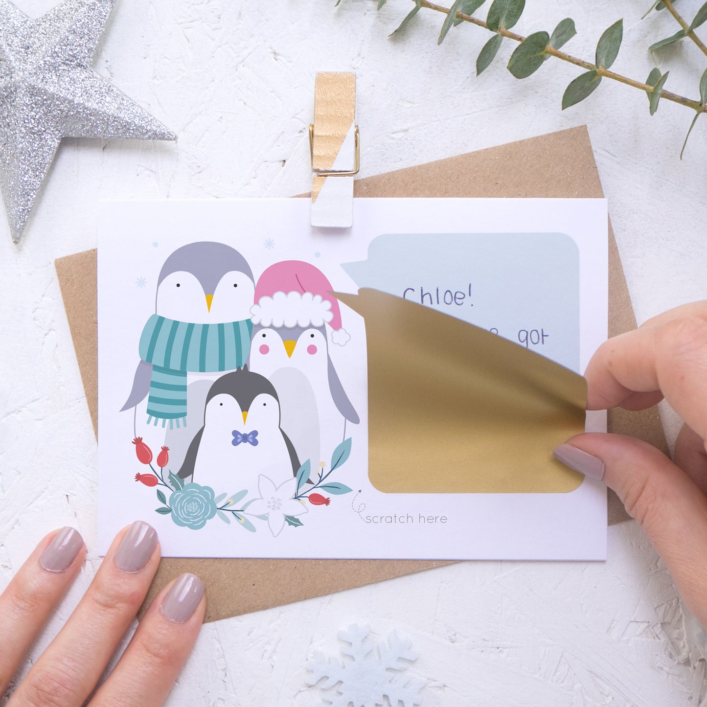 A personalised penguin family scratch card where the sticking down of the gold scratch panel is being demonstrated. Shot on a white background with a glittery star and sprig of eucalyptus.