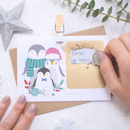 A personalised penguin family scratch card where the scratching off of the gold panel is being demonstrated. Shot on a white background with a glittery star and sprig of eucalyptus.