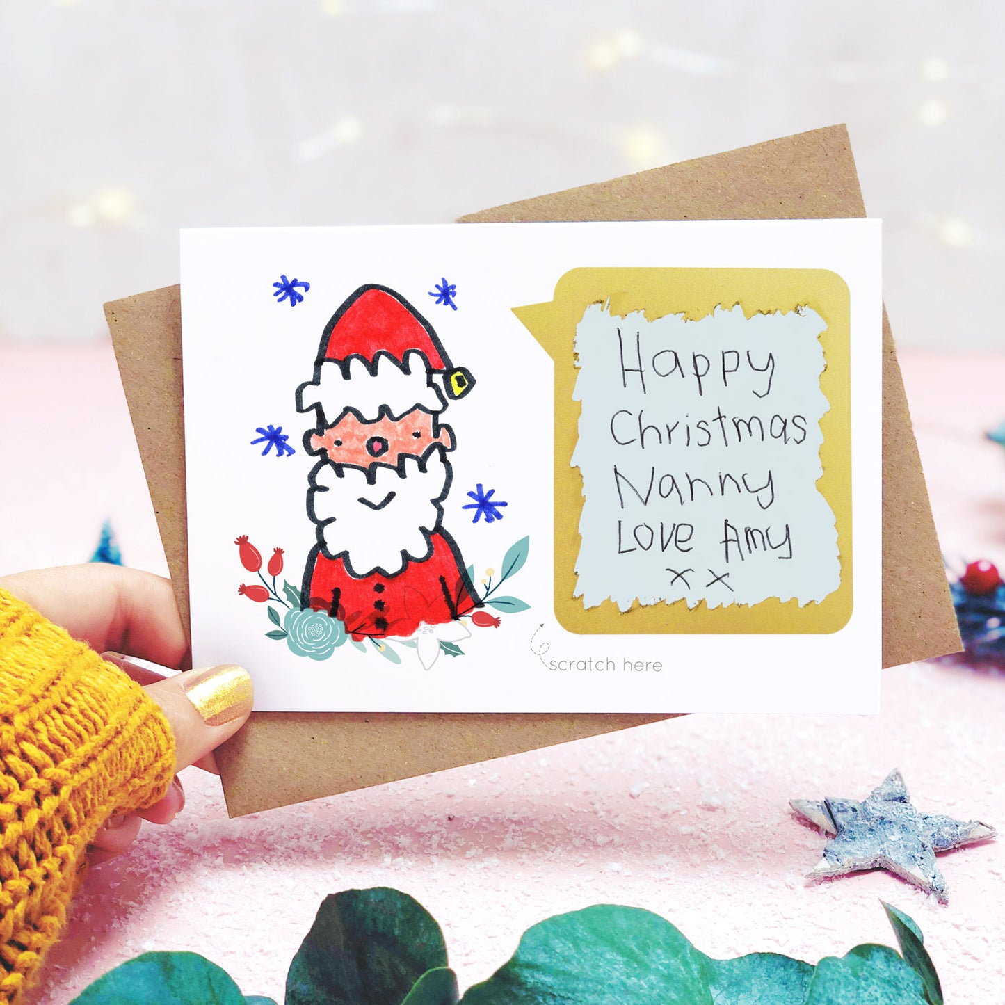 A make your own Christmas scratch card showing a childs drawing of father christmas and the scratch panel having been scratched to reveal the hand written message. Shot on pink and white with a hand and greenery.
