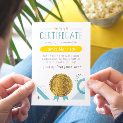 A personalised fathers day certificate printed on white card with varying tones of blue and pops of yellow and gold. Each certificate has a shiny gold seal and space to sign your name. Photographed over a lap with a yellow foot rest in the background.