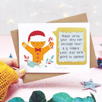 A personalised gingerbread man scratch card shot in a lifestyle setting with a pink background being held behind a sprig of eucalyptus and festive props. The scratch panel has been scratched off to reveal the hidden message.