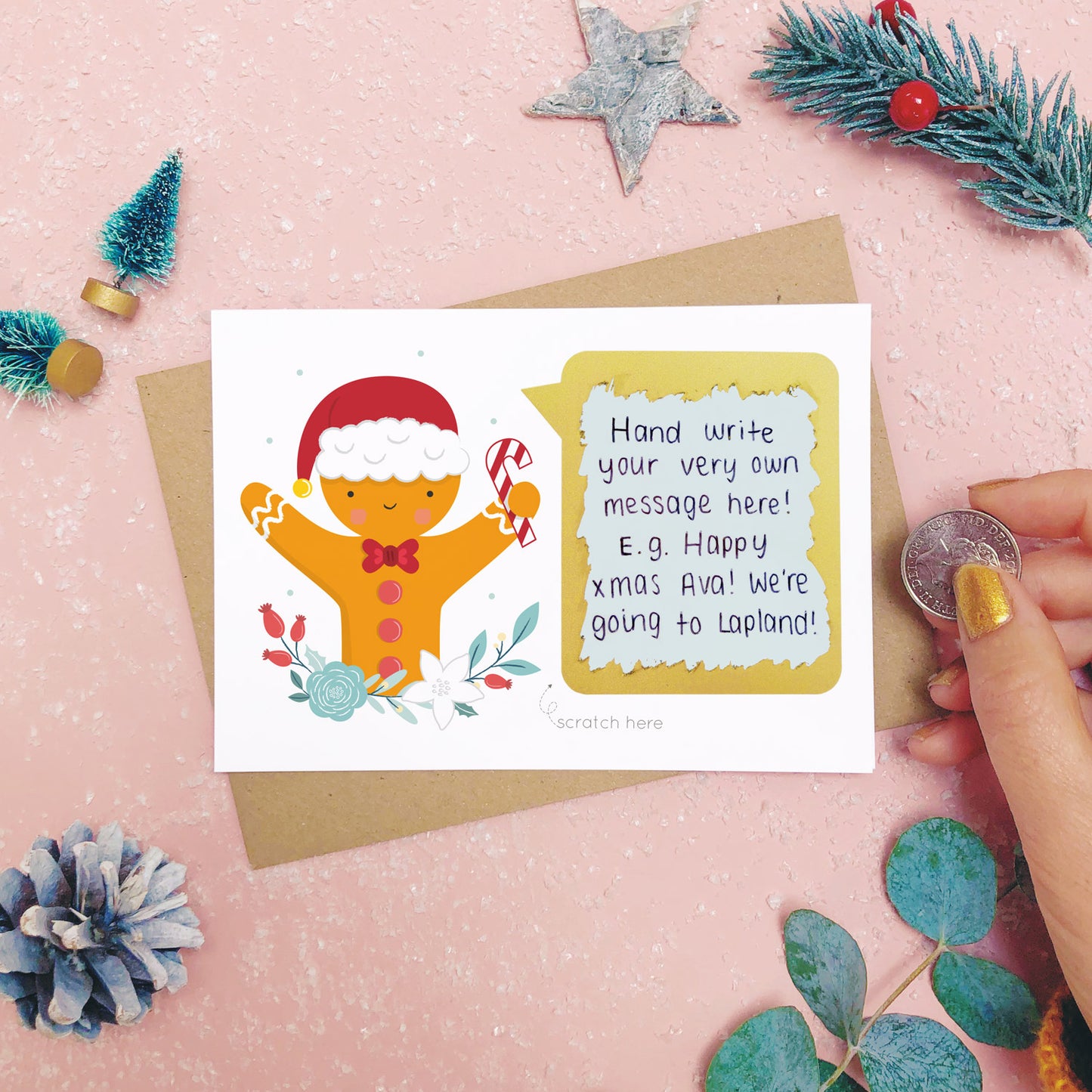 A personalised gingerbread man scratch card after the gold panel has been attached.Shot on a pink background with grey and green festive props.