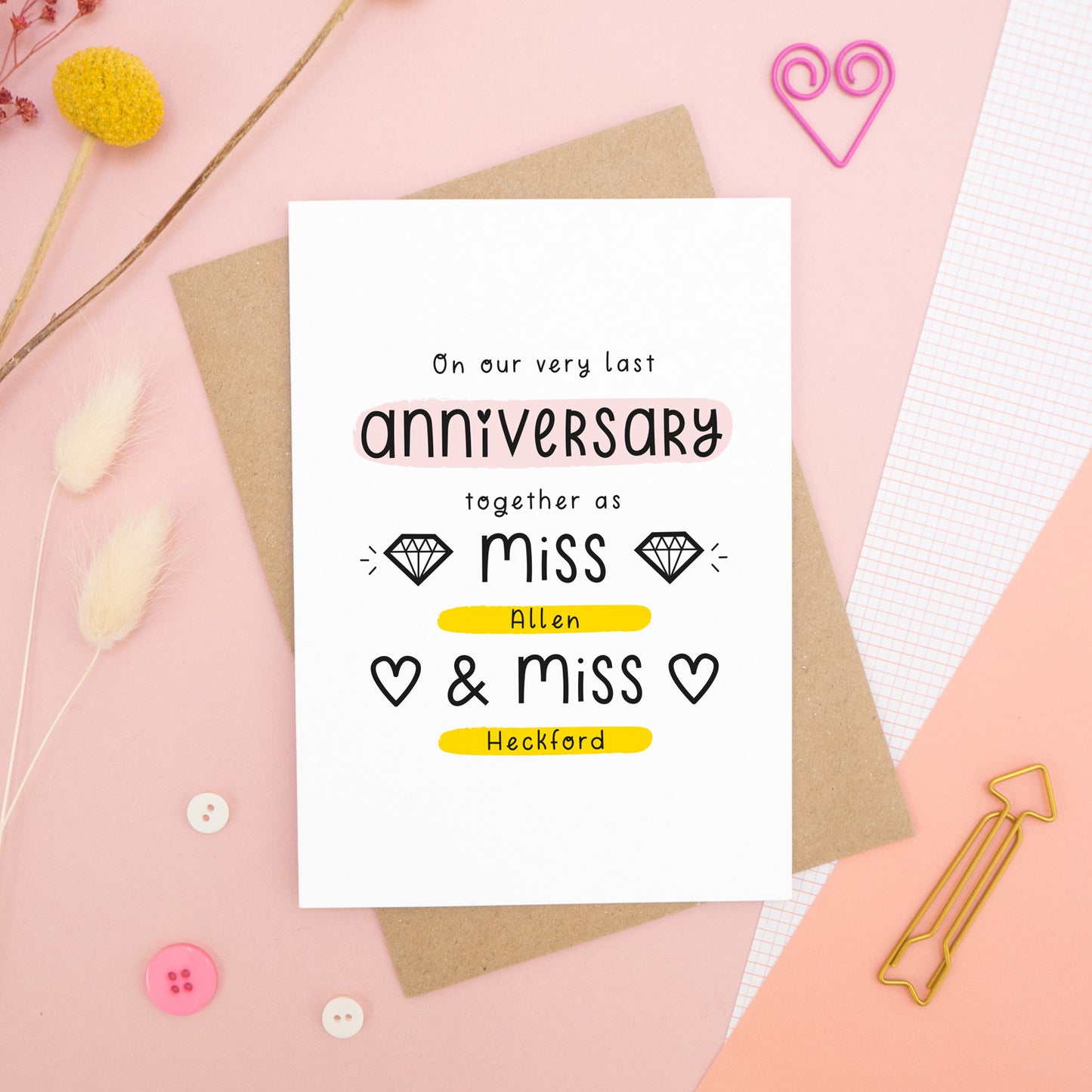 A personalised last anniversary or Valentine’s card photographed on a pink background with floral props, paper clips, and buttons. This image shows the last anniversary option with the Miss & Miss wording. The text is black and there are pops of yellow and pink behind key words.