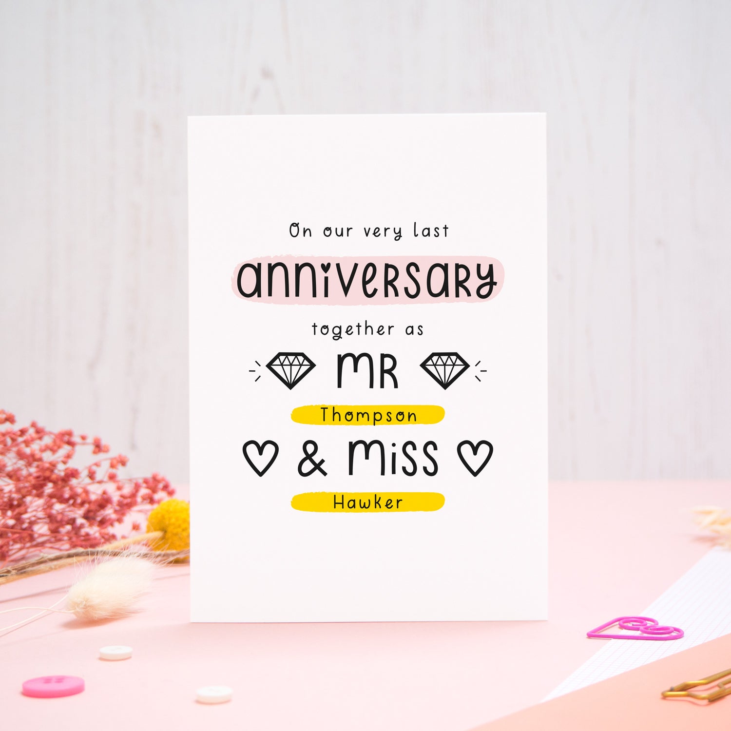 A personalised last anniversary or Valentine’s card photographed on a pink and white background with floral props, paper clips, and buttons. This image shows the last anniversary option with the Mr & Miss wording. The text is black and there are pops of yellow and pink behind key words.