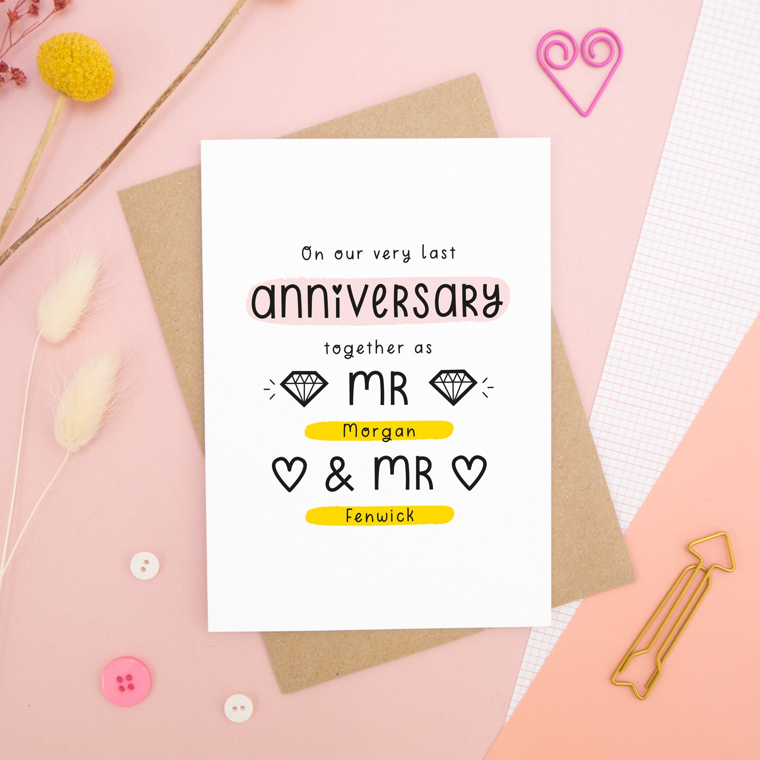 A personalised last anniversary or Valentine’s card photographed on a pink background with floral props, paper clips, and buttons. This image shows the last anniversary option with the Mr & Mr wording. The text is black and there are pops of yellow and pink behind key words.