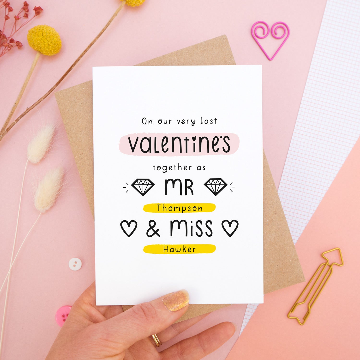 A personalised last anniversary or Valentine’s card photographed on a pink background with floral props, paper clips, and buttons. This image shows the last valentine’s option with the Mr & Miss wording. The text is black and there are pops of yellow and pink behind key words.