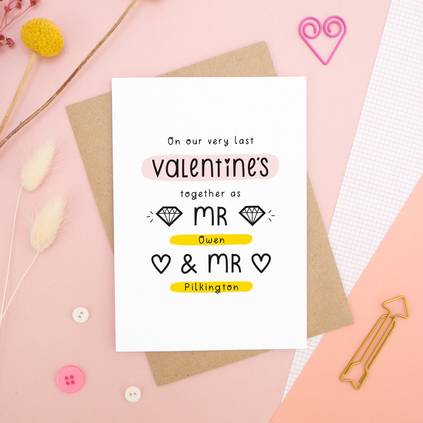 A personalised last anniversary or Valentine’s card photographed on a pink background with floral props, paper clips, and buttons. This image shows the last valentine’s option with the Mr & Mr wording. The text is black and there are pops of yellow and pink behind key words.