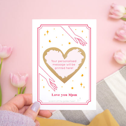 A personalised 'love you...' scratch card with the gold heart scratched off revealing where the personalised message will be printed. The card is photographed held over a pink background with tulips!