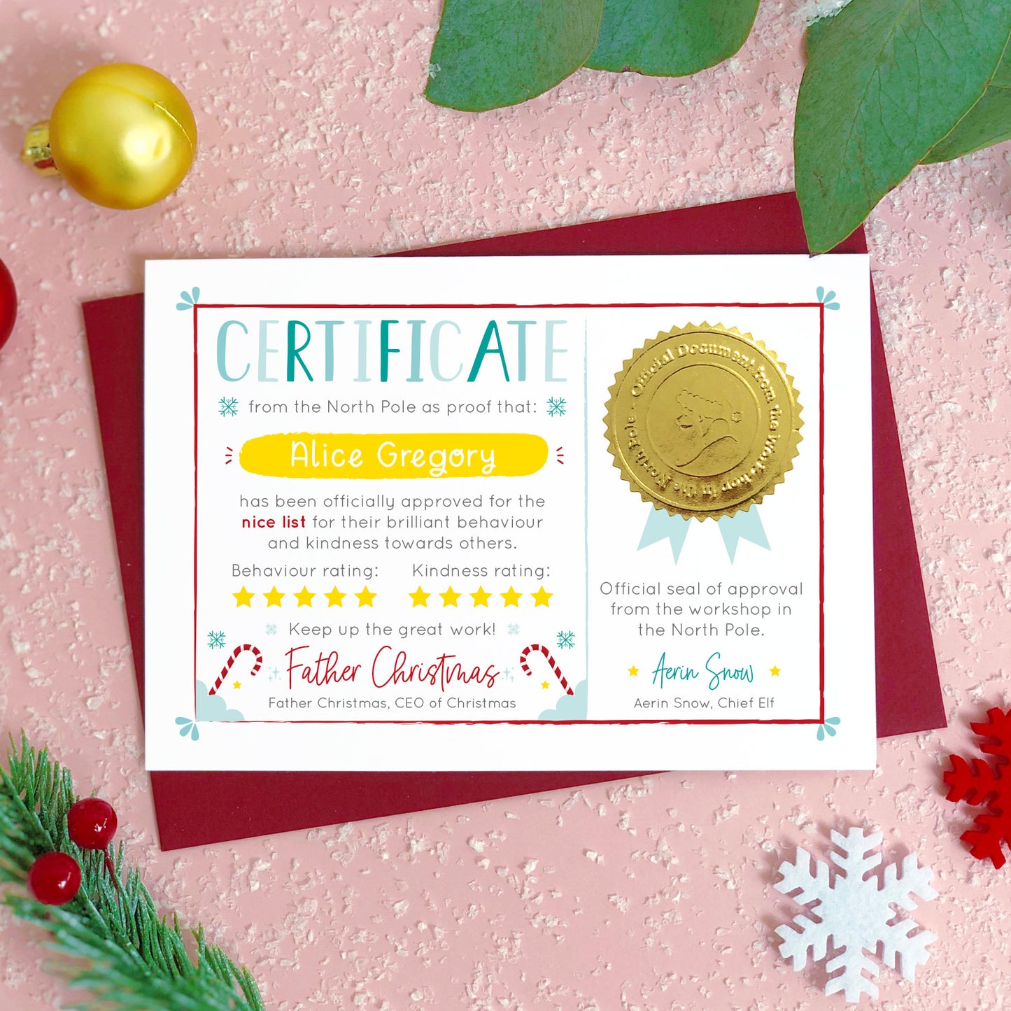 A personalised nice list certificate card from Father Christmas featuring a shiny gold seal, and signatures from santa and his chief elf. Shot on a pink background with baubles, snowflakes and Christmas foliage.
