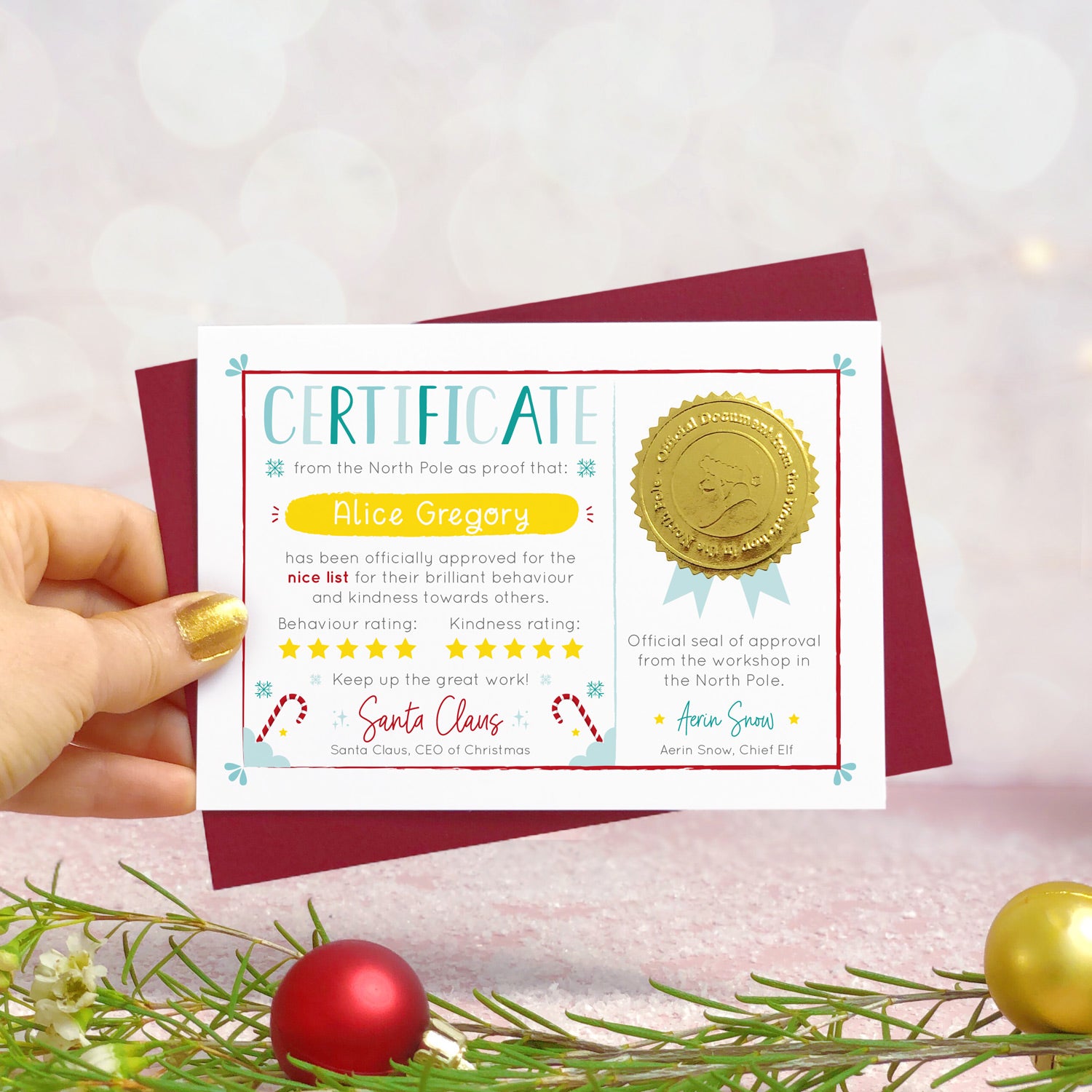 A personalised nice list certificate card from Santa Claus featuring a shiny gold seal, and signatures from Father Christmas and his chief elf. Shot on a pink background with baubles, fairy lights and Christmas foliage.