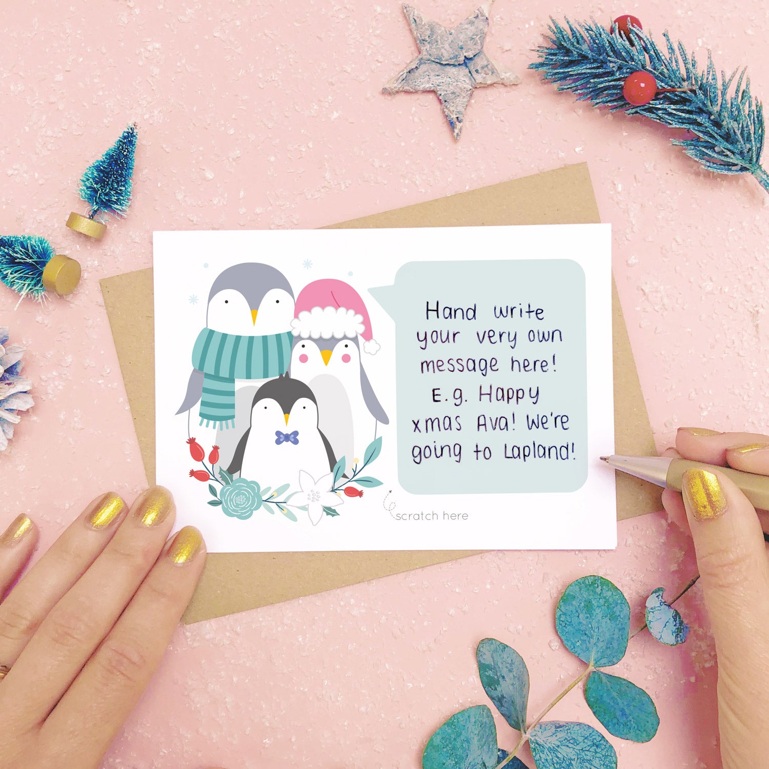 A personalised penguin family scratch card showing where to write the hand written message. Shot on a pink background with grey and green festive props.