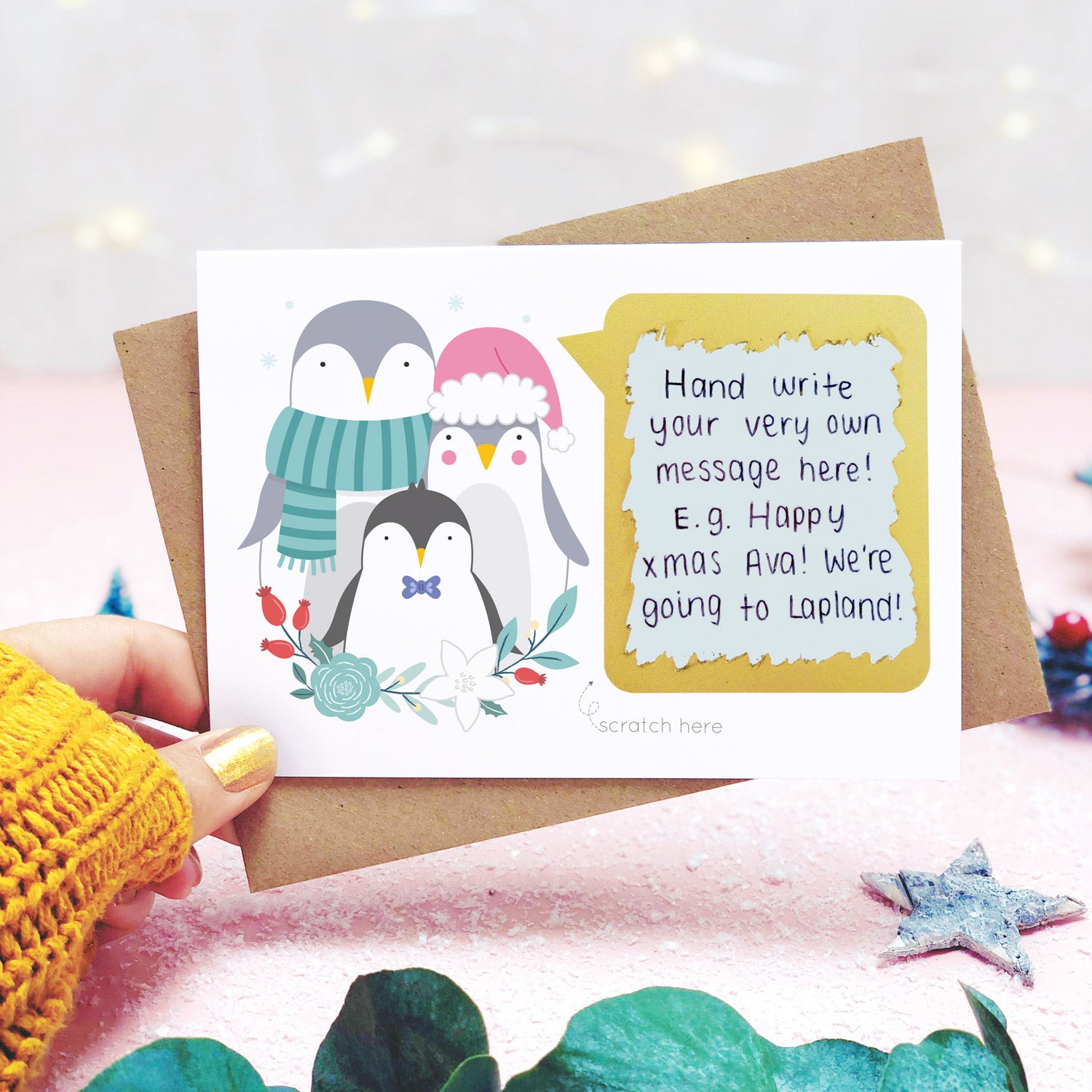 A personalised penguin family scratch card shot in a lifestyle setting with a pink background being held behind a sprig of eucalyptus and festive props. The scratch panel has been scratched off to reveal the hidden message.