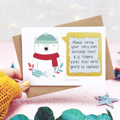 A personalised polar bear scratch card with the hand written message revealed. Shot on a pink background, behind a sprig of eucalyptus and festive props.