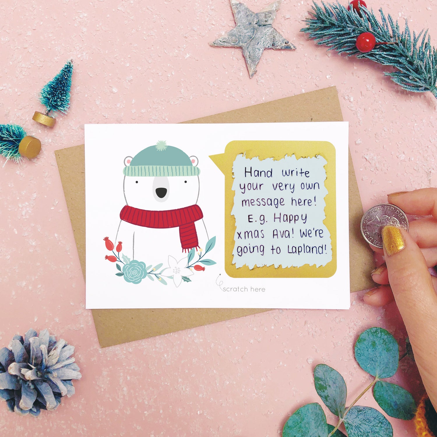A personalised polar bear scratch card shot on a pink background with festive props in grey and green. The scratch panel has been scratched off with a coin to reveal the hidden message.