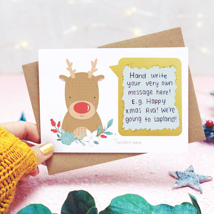 A personalised red nose reindeer scratch card shot in a lifestyle setting with a pink background being held behind a sprig of eucalyptus and festive props. The scratch panel has been scratched off to reveal the hidden message.