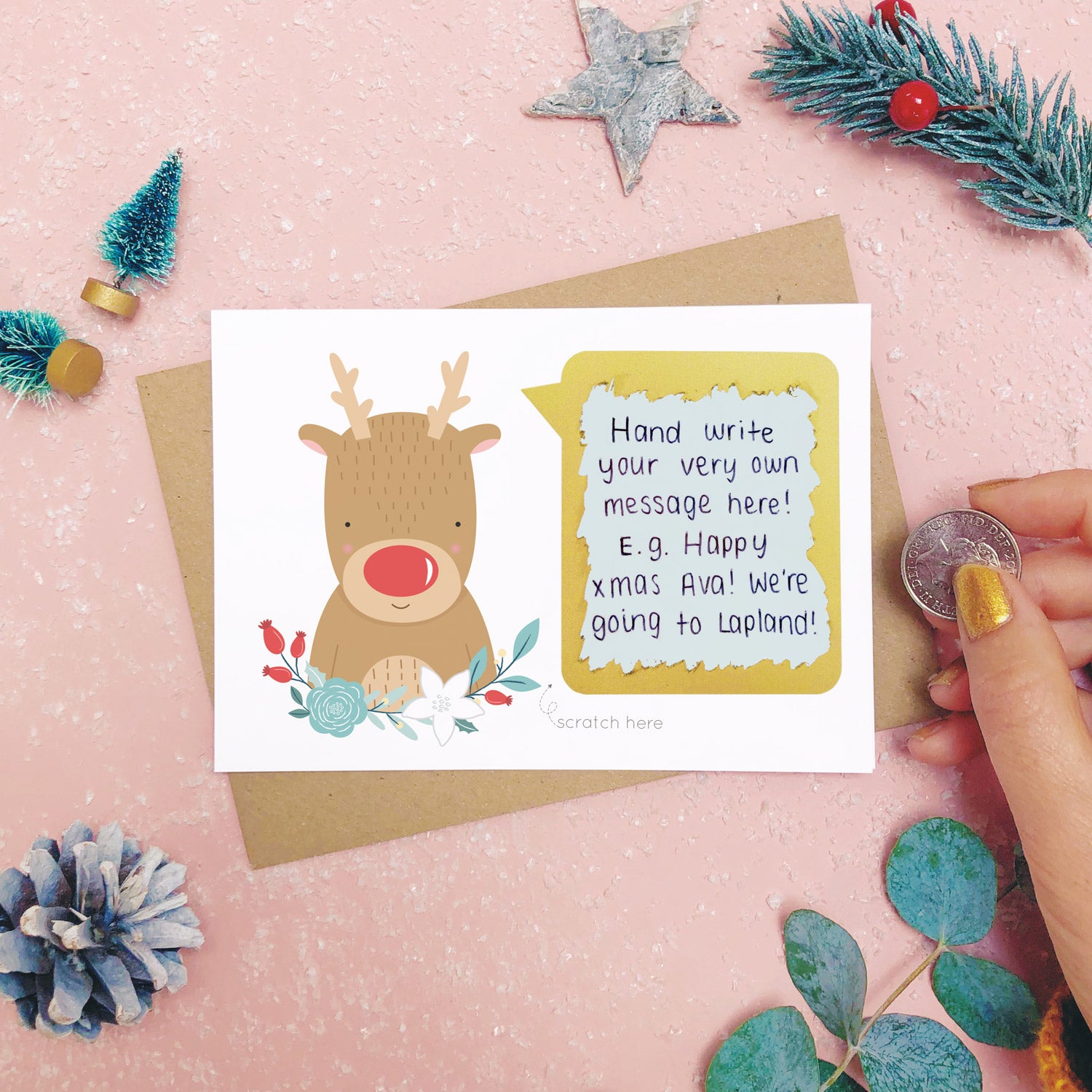 A personalised red nose reindeer scratch card after the scratch panel has been scratched with a coin to reveal the hidden message.Shot on a pink background with grey and green festive props.