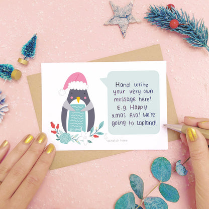 Santa Penguin scratch card showing where you write your hidden message. Shot on a pink background, surrounded with festive christmas props in tones of green and grey.