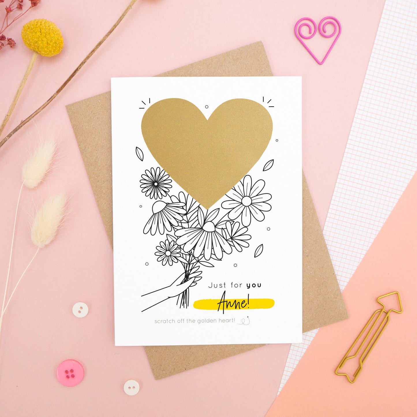 A personalised flower bouquet scratch card photographed on a pink background with floral props, paper clips, and buttons. This card shows a black and white floral illustration and the golden heart before it is scratched off to reveal the secret message!