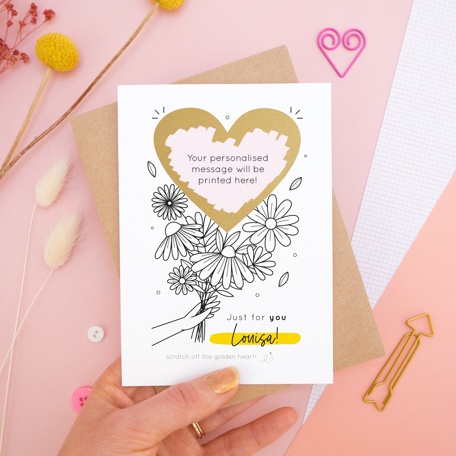 A personalised flower bouquet scratch card photographed on a pink background with floral props, paper clips, and buttons. This card shows a black and white floral illustration and the golden heart after it has been scratched off to reveal the secret message!