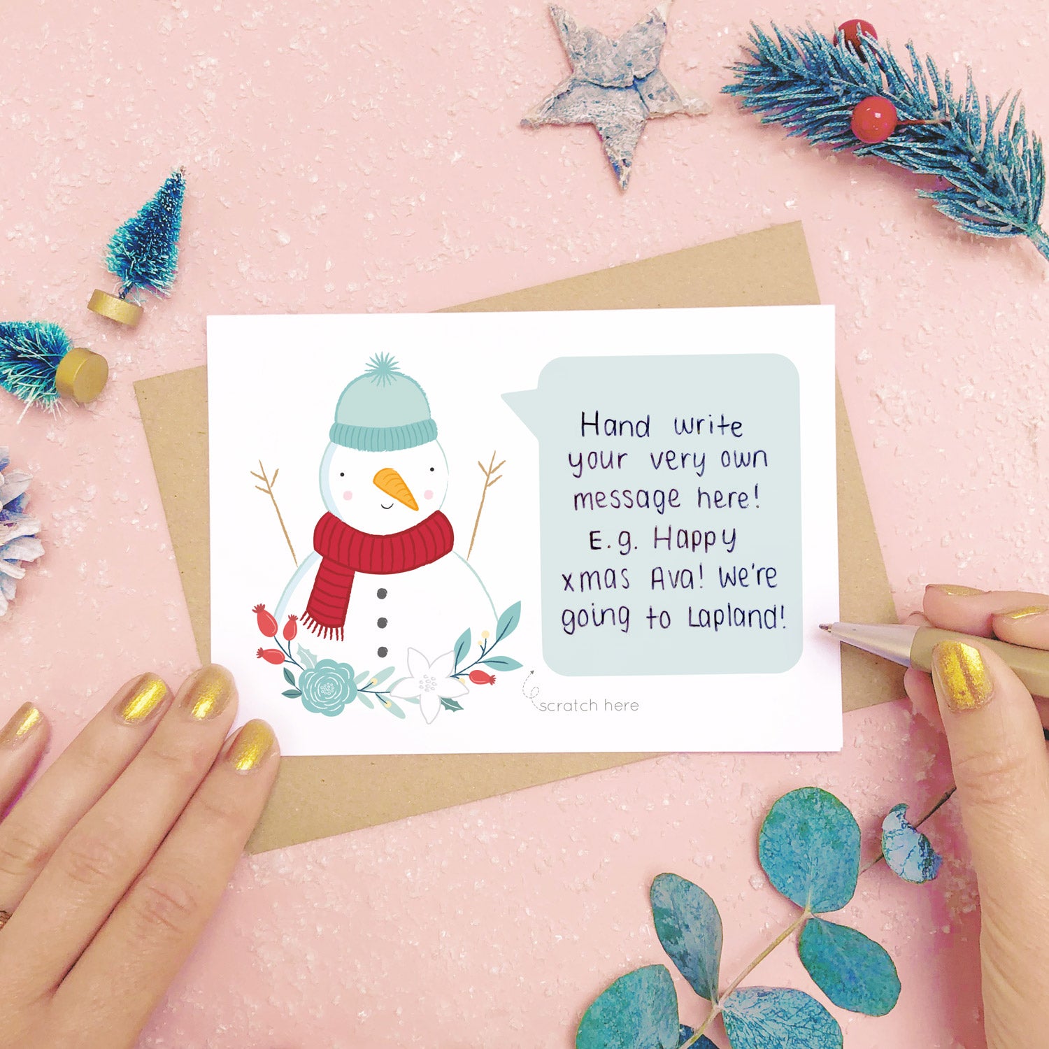 A personalised snowman scratch card showing where to write the hand written message. Shot on a pink background with grey and green festive props.