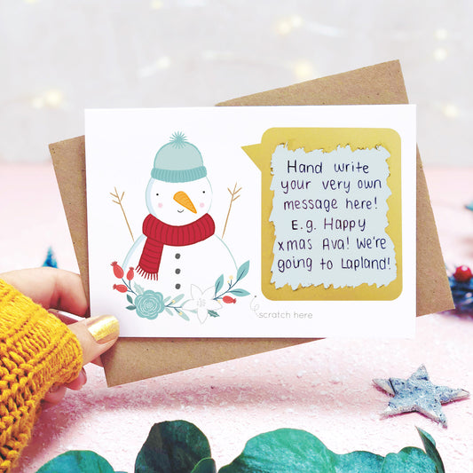 A personalised snowman scratch card shot in a lifestyle setting with a pink background being held behind a sprig of eucalyptus and festive props. The scratch panel has been scratched off to reveal the hidden message.