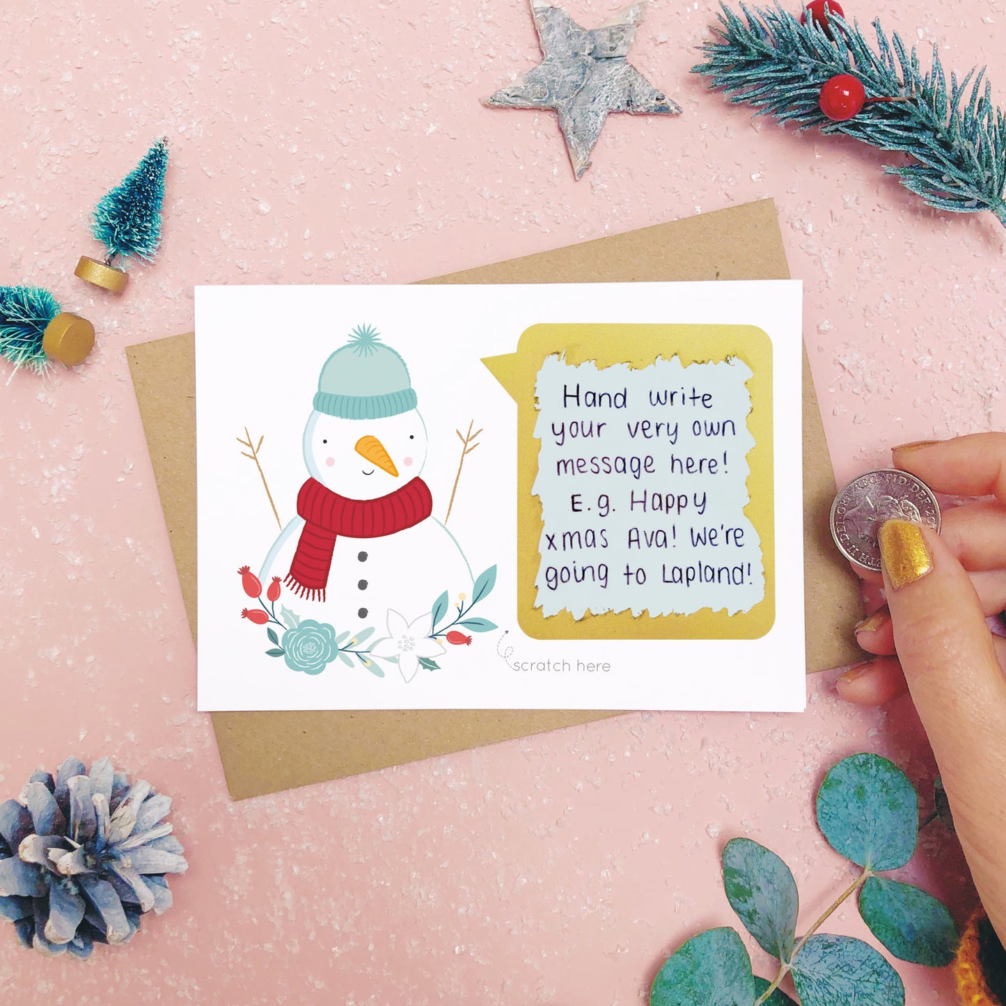 A personalised snowman scratch card after the scratch panel has been scratched with a coin to reveal the hidden message.Shot on a pink background with grey and green festive props.