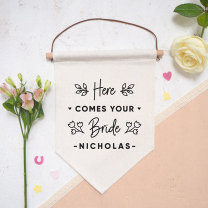 A linen pennant flag with the words 'here comes your bride [insert name of groom]. Each flag features leafy vines and little hearts. This flag has been photographed on a white and peach background with confetti and flowers either side.